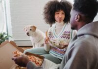 Crop African American couple speaking with each other while having snack with pizza and salad at home with hungry dog